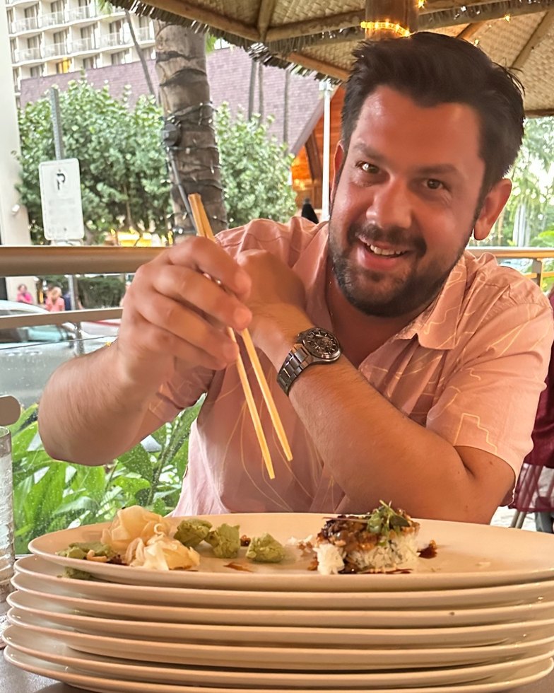 Panagiotis at an outdoor restaurant in a pink shirt, grinning, holding chopsticks above a stack of plates